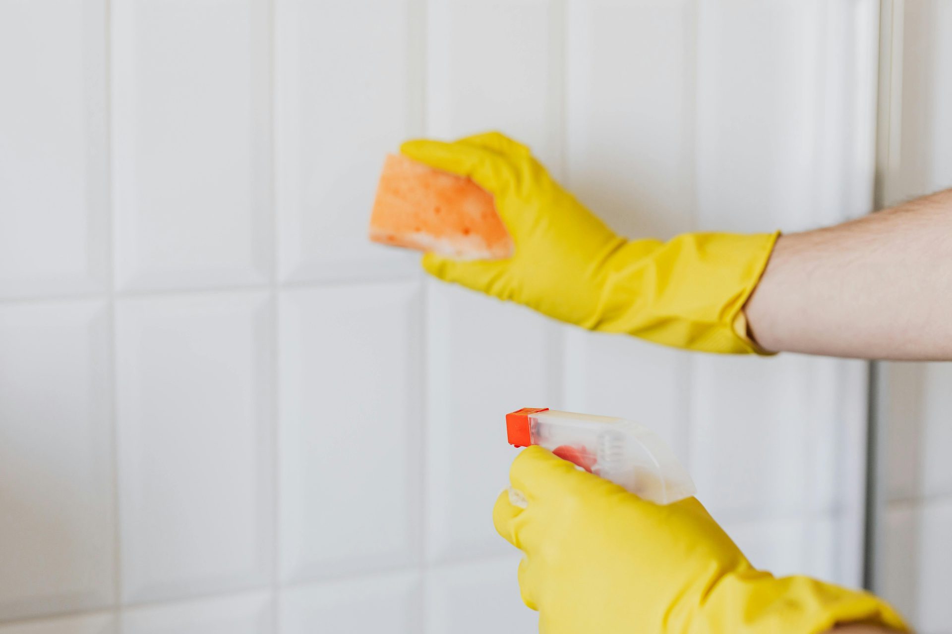 Une personne portant des gants jaunes nettoie les carreaux d'une salle de bain avec une éponge et un vaporisateur.
