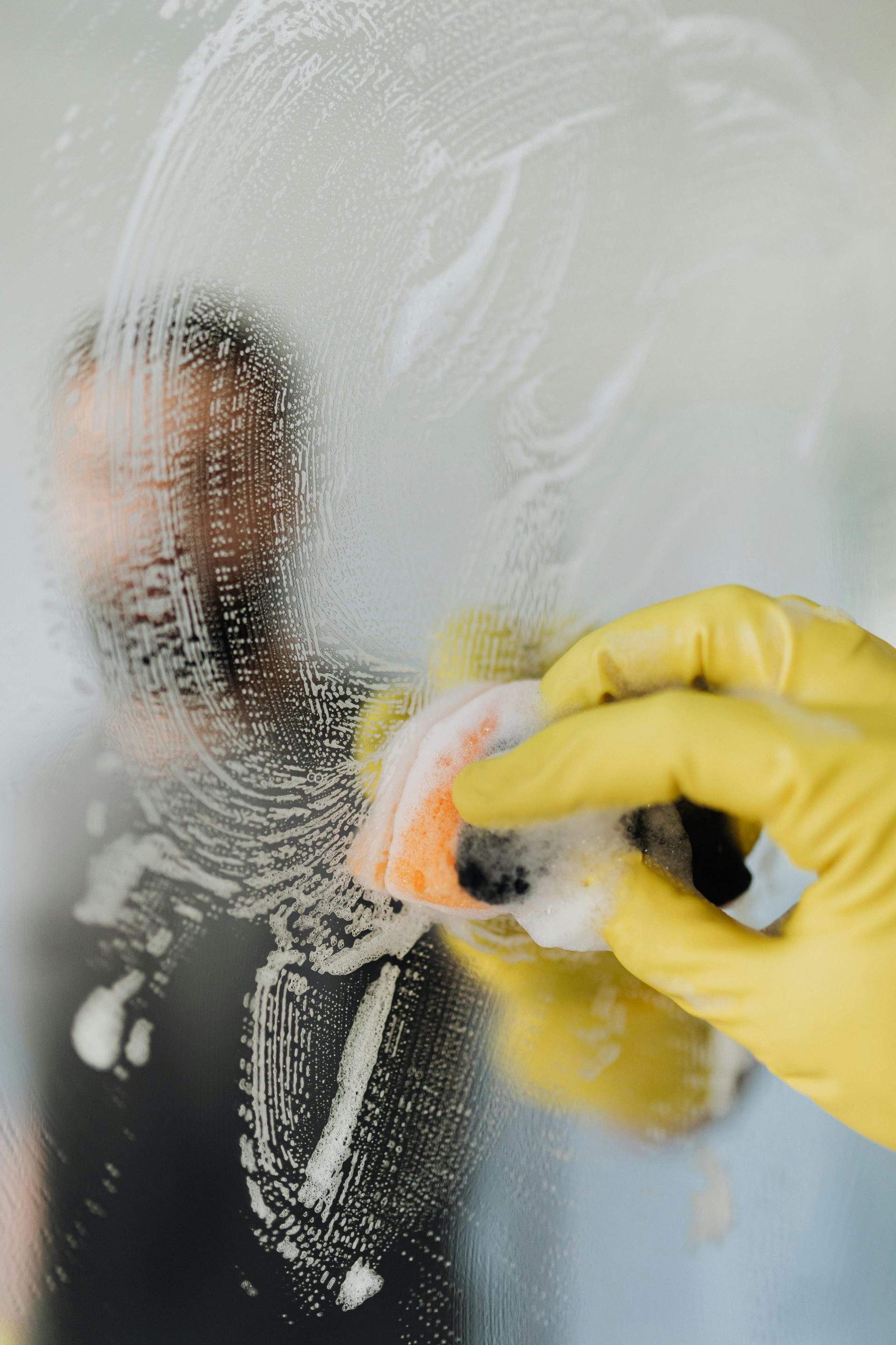 Un homme portant des gants jaunes nettoie un miroir de salle de bains avec une éponge savonneuse, garantissant ainsi l'hygiène et la propreté.